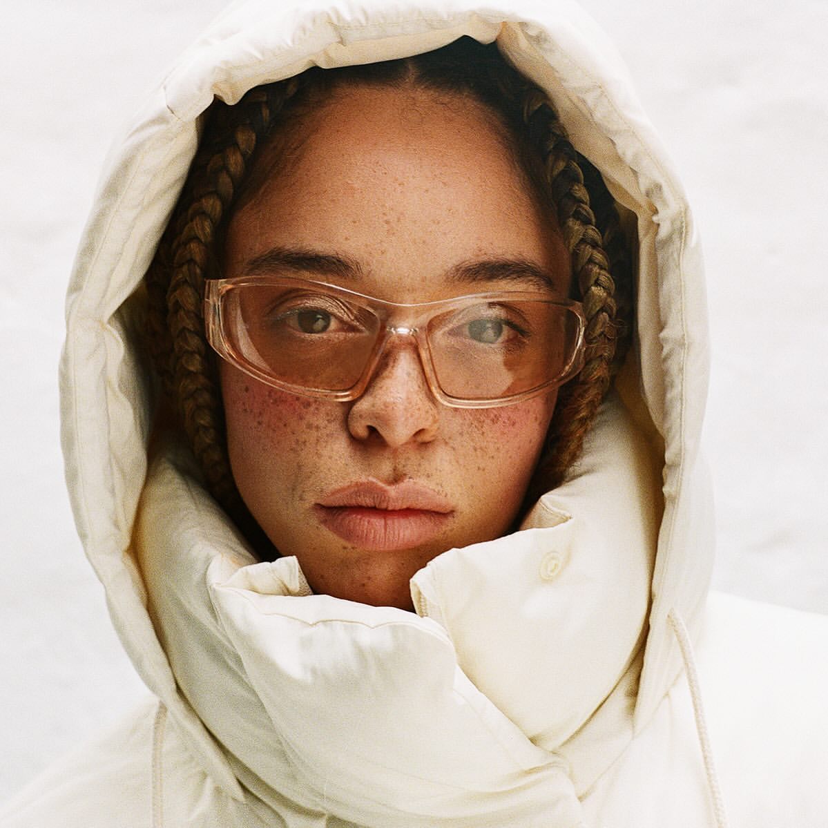 Close-up portrait of a woman with freckles wearing clear sunglasses and a padded hooded jacket, against a neutral background.