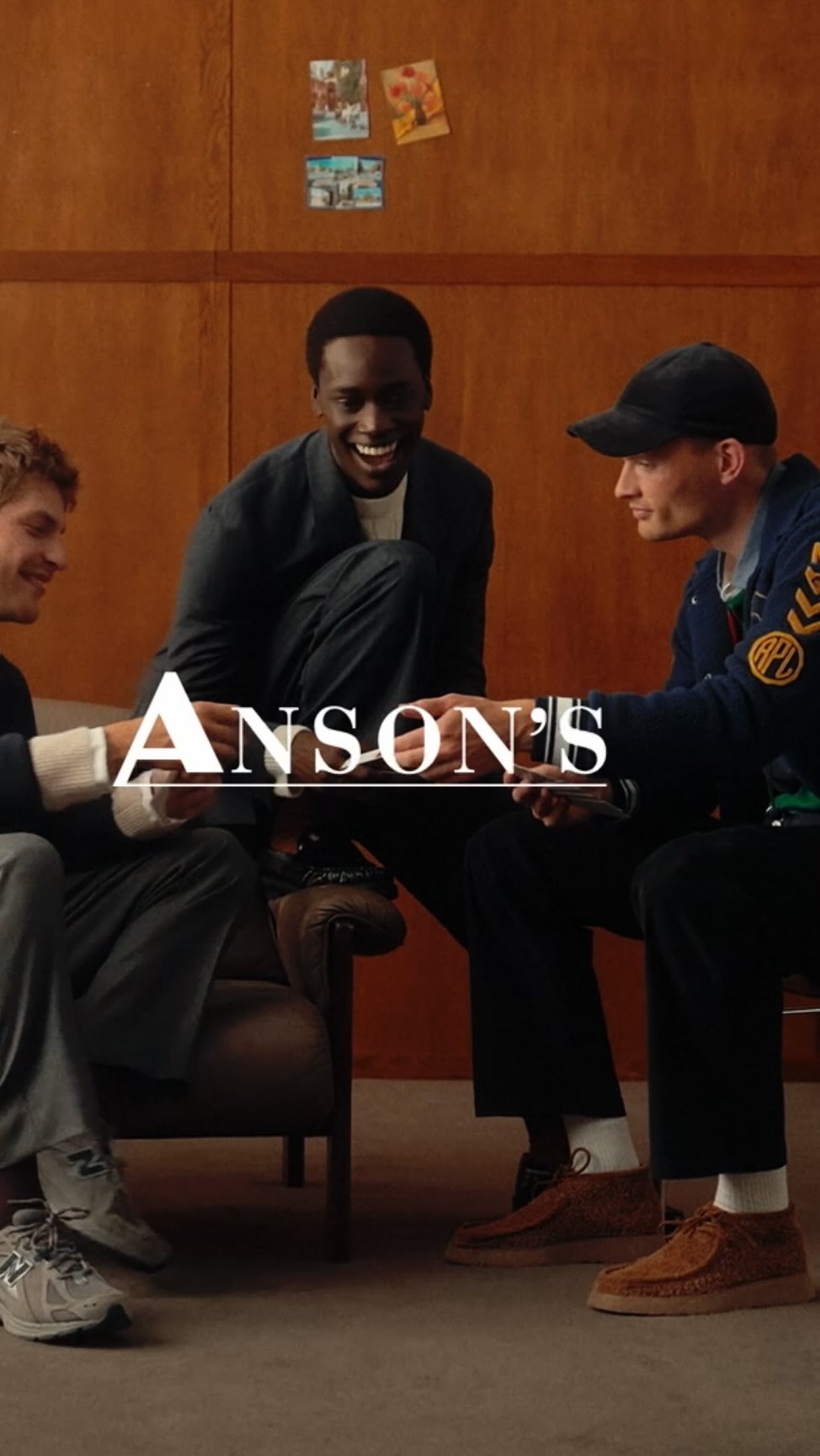 Three stylishly dressed men sit indoors, possibly in a lounge or waiting area. They're engaged in conversation, creating a relaxed scene.