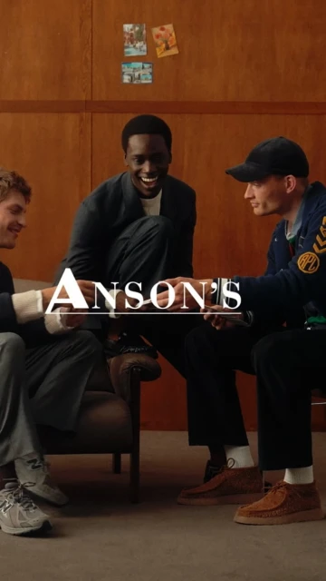 Three stylishly dressed men sit indoors, possibly in a lounge or waiting area. They're engaged in conversation, creating a relaxed scene.