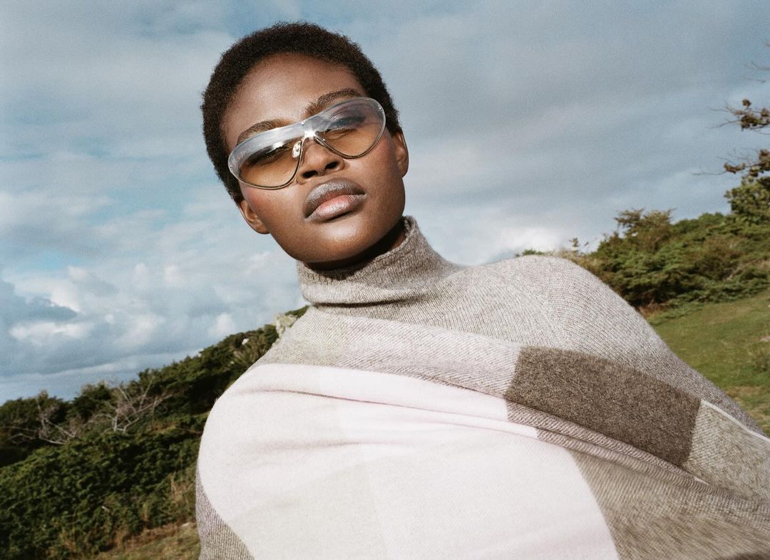 Woman with short dark hair wears sunglasses, a gray turtleneck, and a large pale plaid scarf against a blue sky and green hillside.