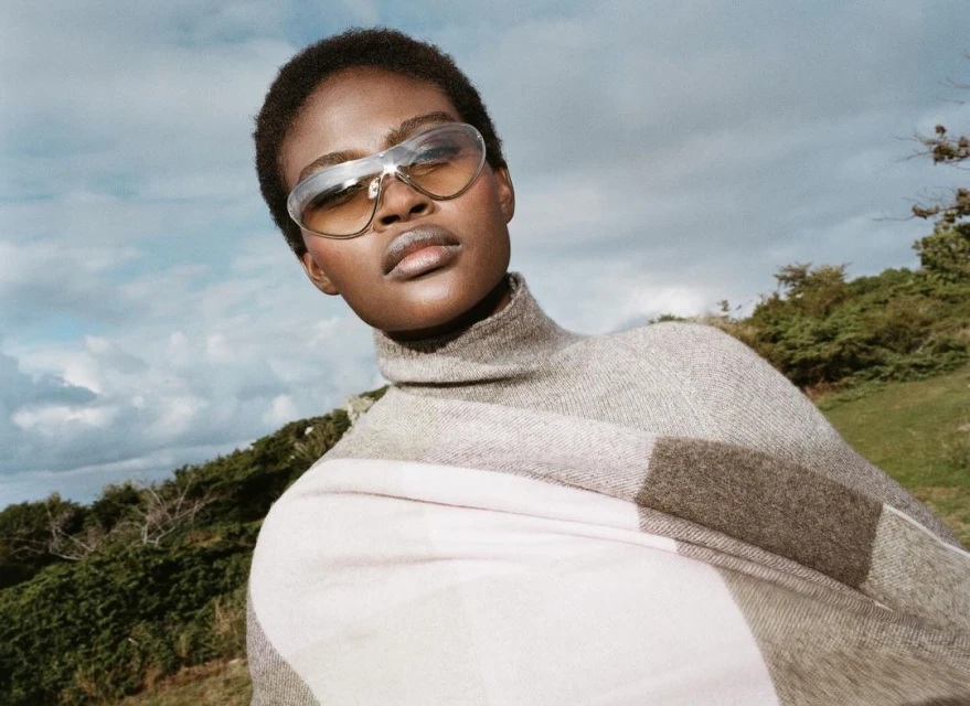 Woman with short dark hair wears sunglasses, a gray turtleneck, and a large pale plaid scarf against a blue sky and green hillside.