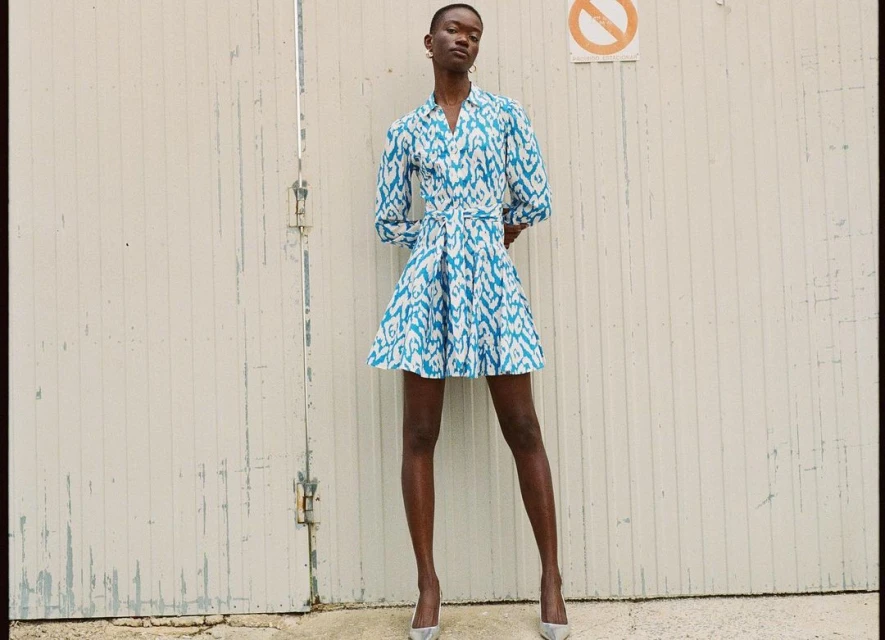 A full-length portrait of a slender Black woman wearing a blue and white patterned dress and silver heels against a white wall.