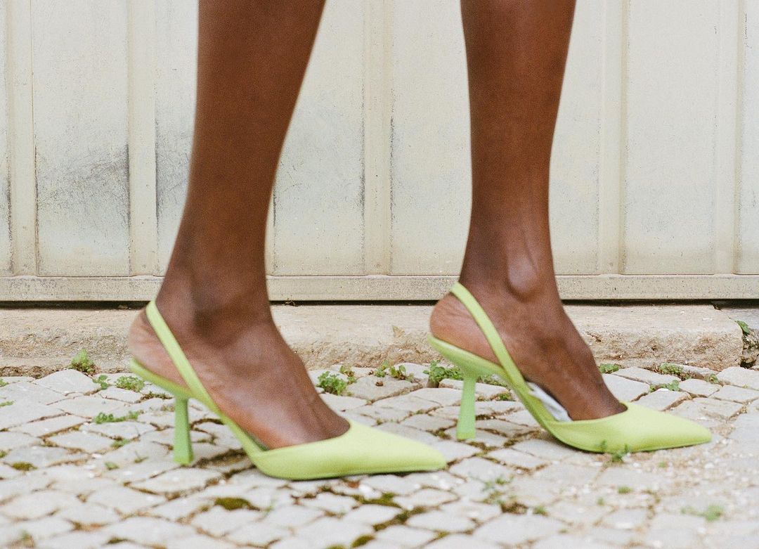 Close-up of an adult female model wearing light green slingback high heel shoes outdoors on a stone surface with a white door behind.
