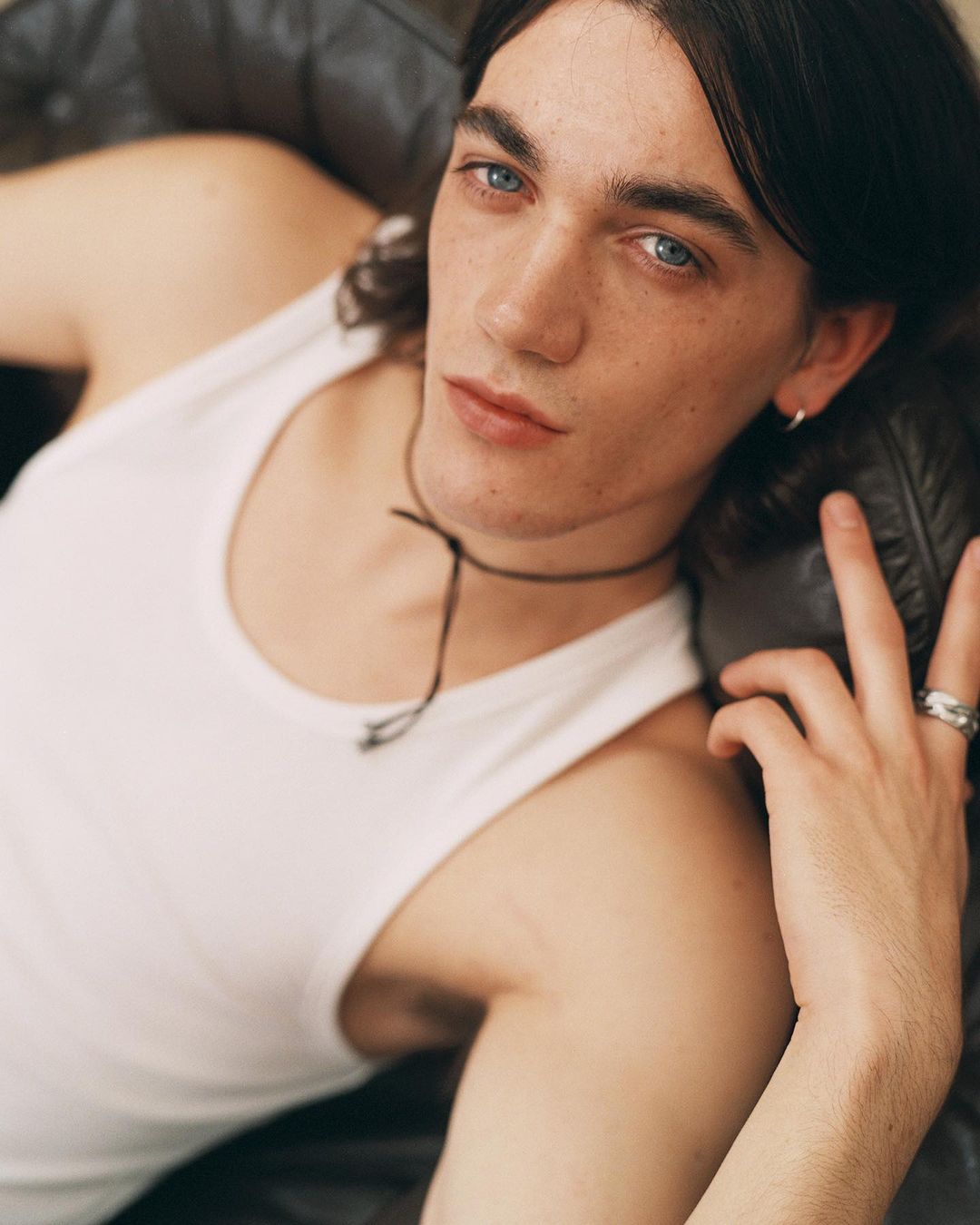 Portrait of a young adult male with long hair, lying down wearing a tank top, silver ring, and a black necklace.