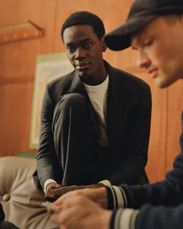 Two men playing cards indoors. One man is Black wearing a jacket and sweater, and the other man is White wearing a baseball cap.