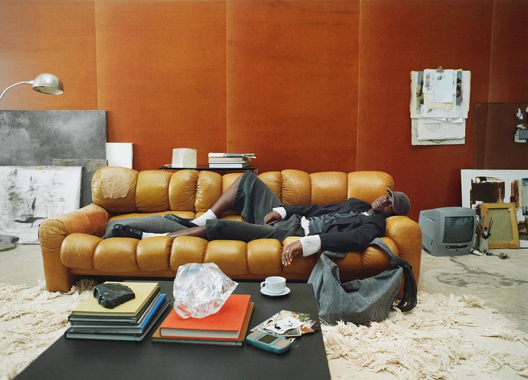An African American man is resting on a brown leather couch with various art objects scattered around the room, creating a relaxed, creative atmosphere.