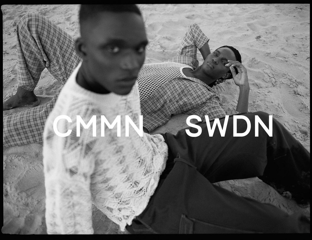 Two dark-skinned men lounge on a sandy beach wearing fashionable summer clothes in a monochrome outdoor fashion editorial.