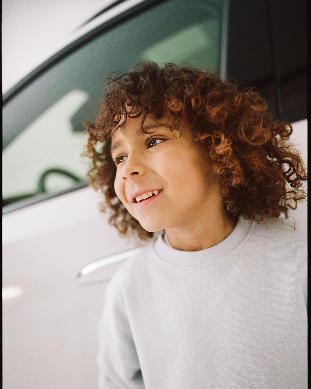 Portrait of a smiling young boy with curly hair standing next to a white car on a sunny day, wearing a light blue sweater.