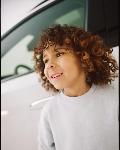 Portrait of a smiling young boy with curly hair standing next to a white car on a sunny day, wearing a light blue sweater.
