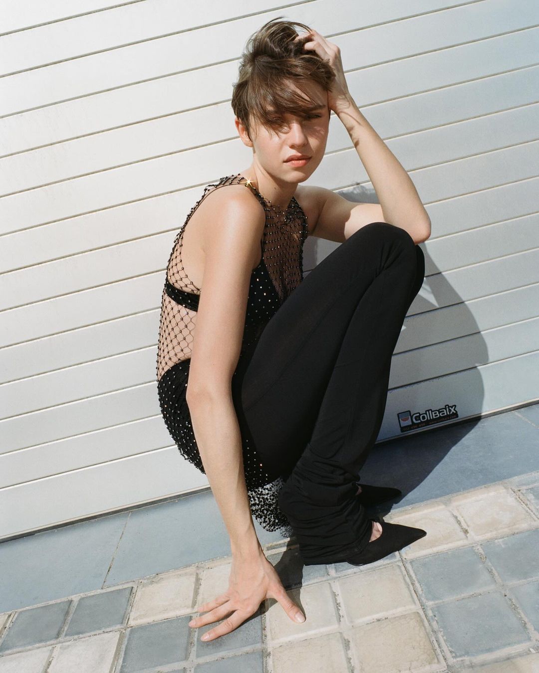 Woman in embellished top and leggings crouching outdoors against a white garage door on a tiled surface, bathed in sunlight.