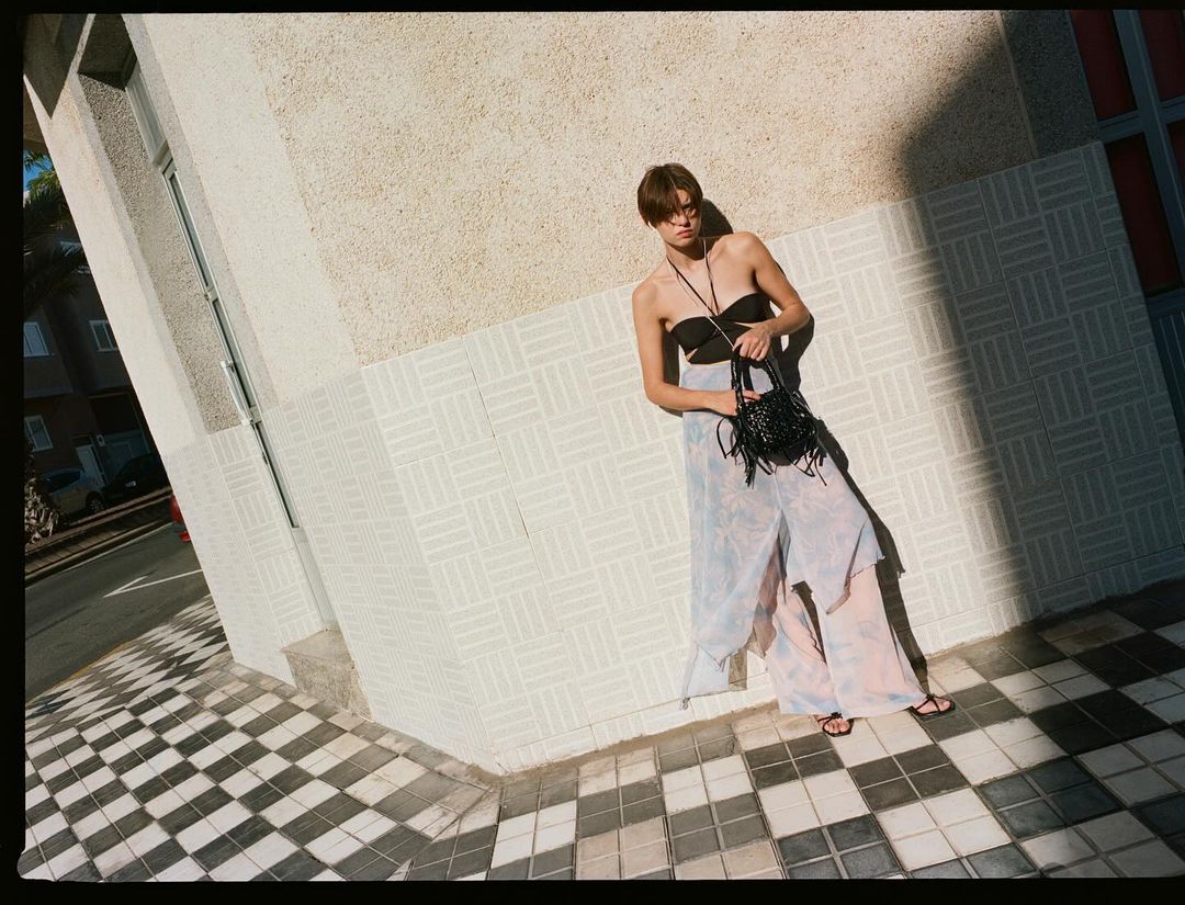 A slim adult fashion model poses against a white tiled wall in a black top and flowing patterned pants, holding a black fringed bag.