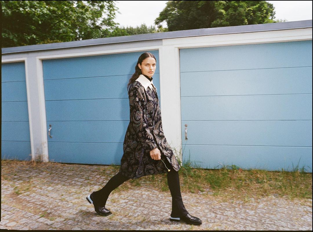 A woman in a patterned coat strides on a paved path next to blue garage doors, a blend of fashion and urban environment.