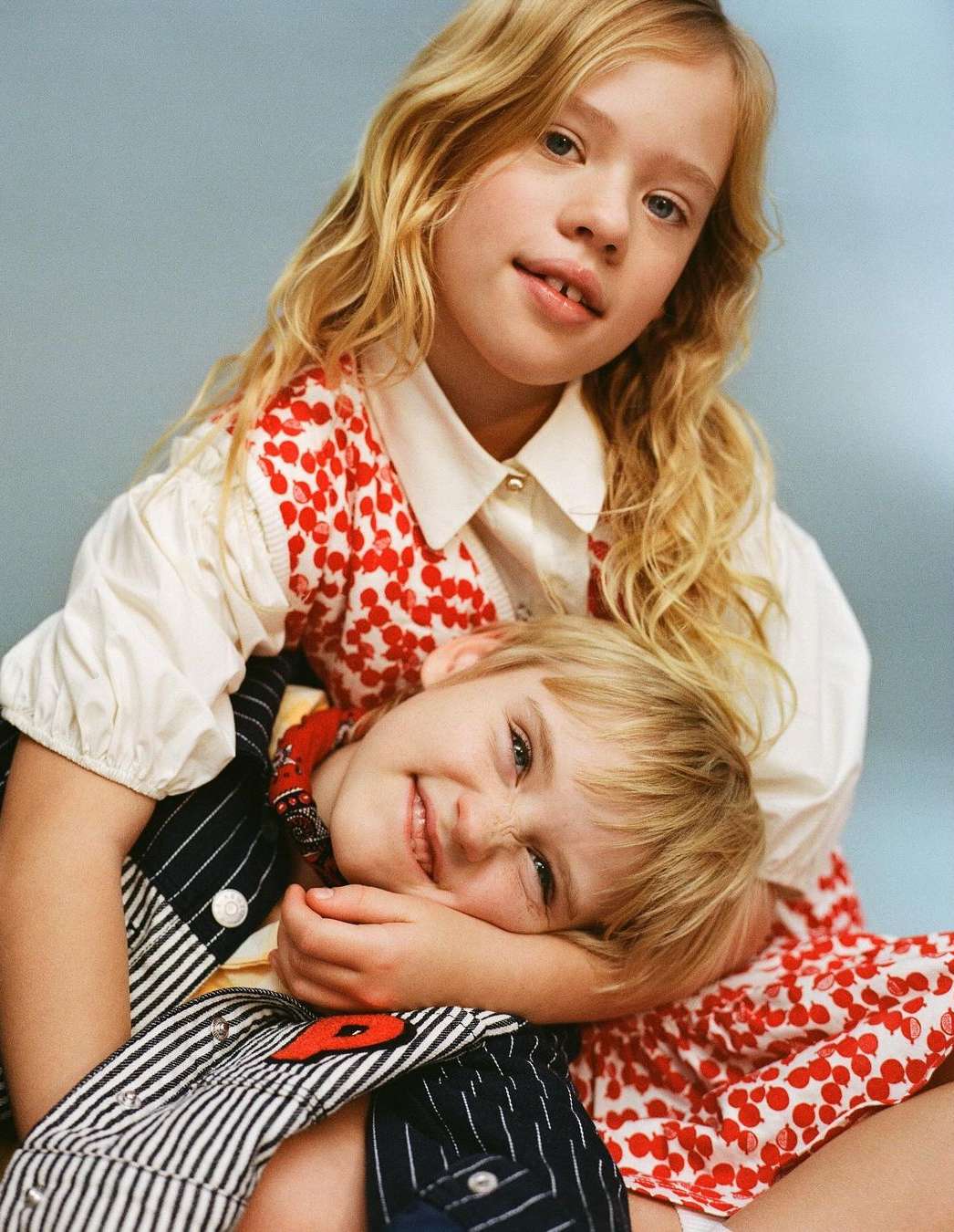 Portrait of a girl with blonde hair embracing a young boy in a photo studio, both smiling. The girl is in a white and red dress.