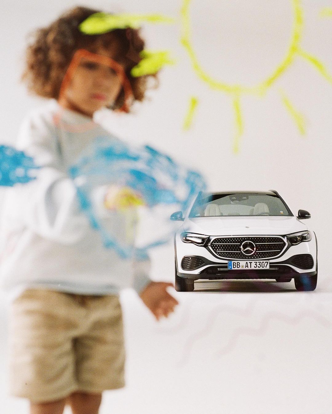 A child stands near a white car in front of a wall with paint. The child is slightly blurred, while the car is in focus.