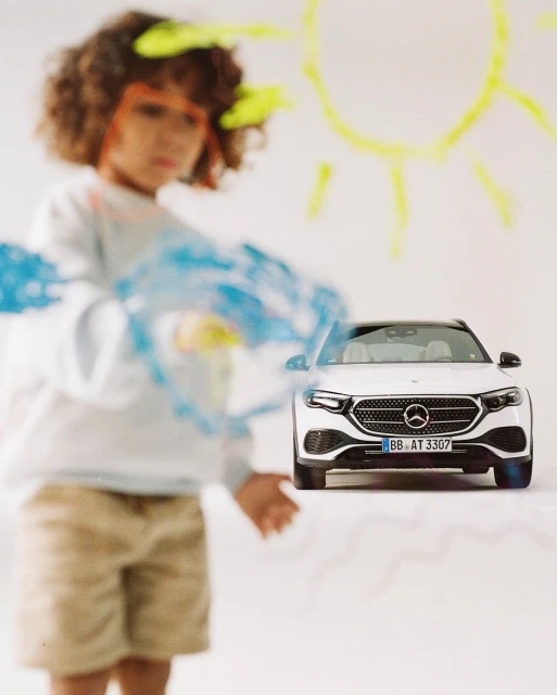 A child stands near a white car in front of a wall with paint. The child is slightly blurred, while the car is in focus.
