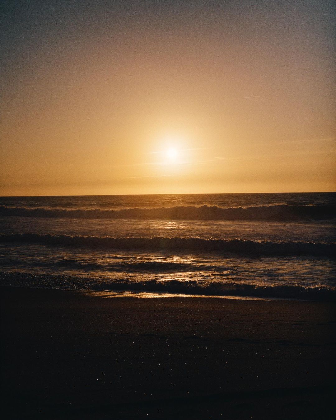 Sunset over the ocean, featuring golden light on the water and waves gently rolling onto a dark sandy beach during golden hour.