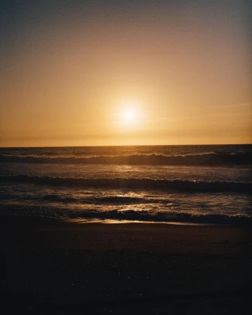 Sunset over the ocean, featuring golden light on the water and waves gently rolling onto a dark sandy beach during golden hour.