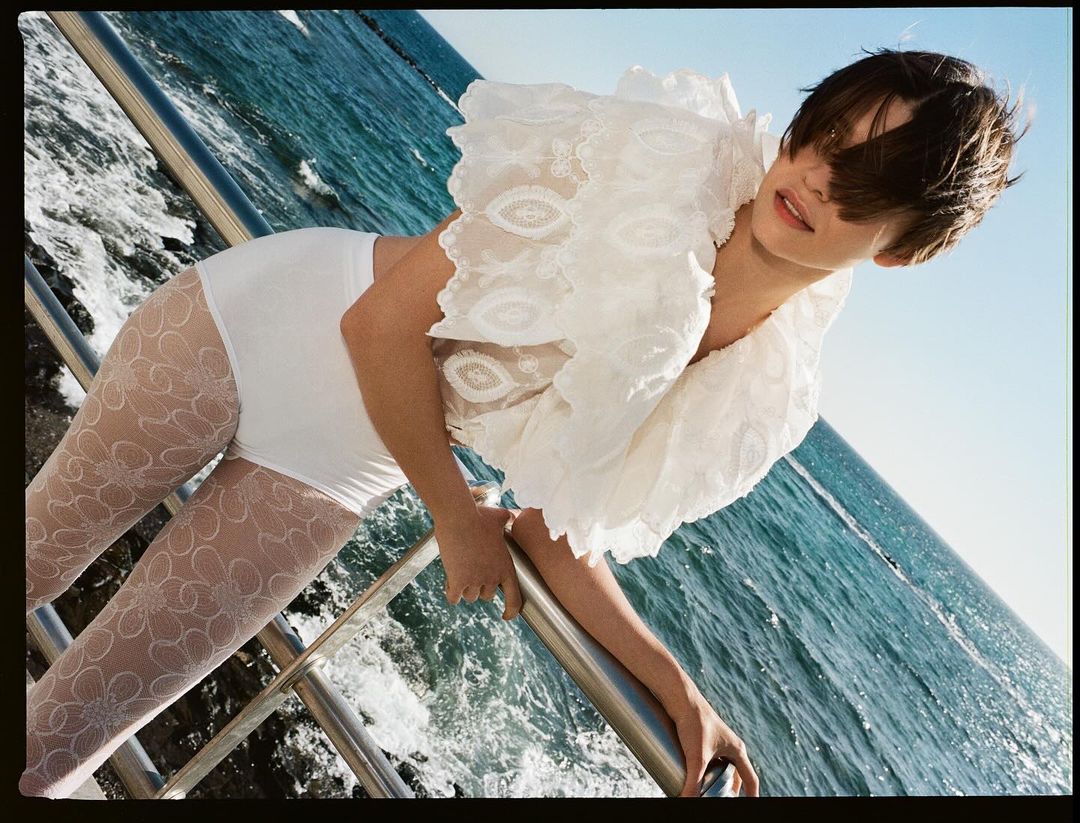 Woman on a boat leaning over the railing wearing a lacy white blouse and patterned tights, in an outdoor, daytime setting with blue water.