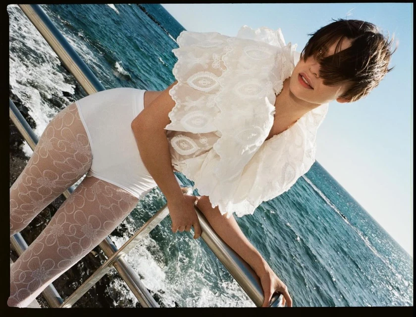 Woman on a boat leaning over the railing wearing a lacy white blouse and patterned tights, in an outdoor, daytime setting with blue water.