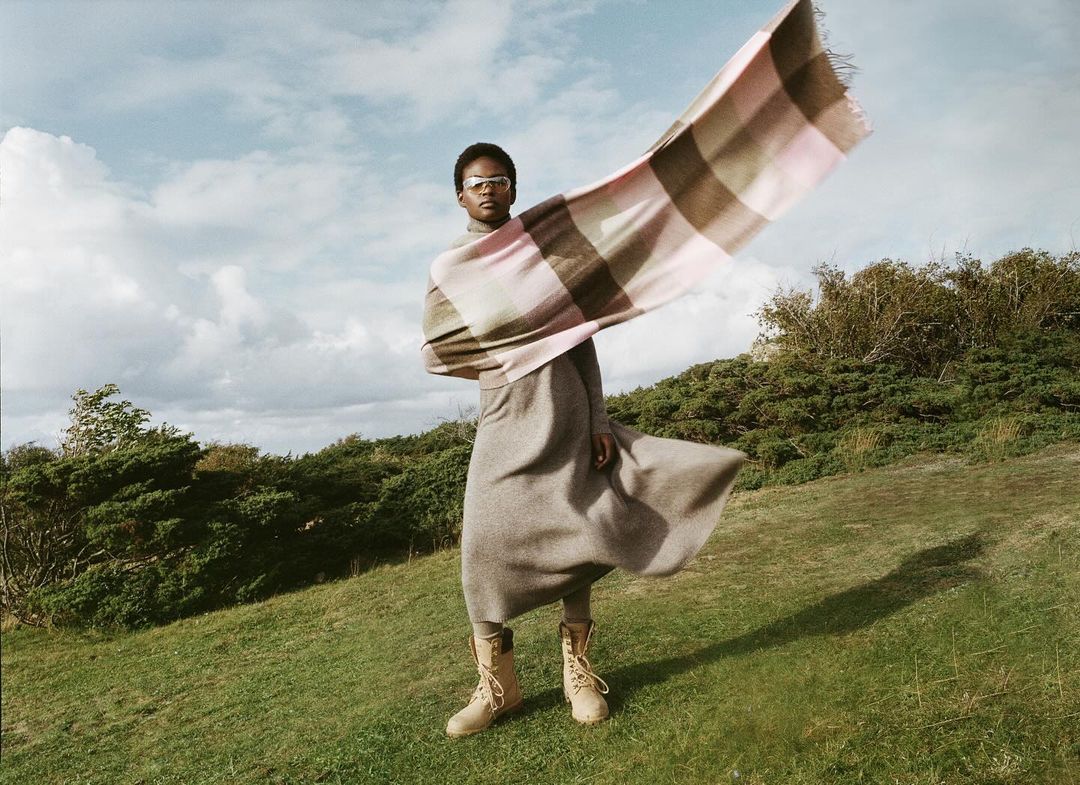 Adult woman wearing tan boots and a long grey dress standing in a grassy field with a pink and beige checkered scarf waving in the wind.