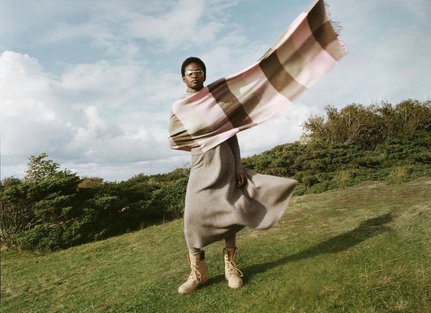 Adult woman wearing tan boots and a long grey dress standing in a grassy field with a pink and beige checkered scarf waving in the wind.