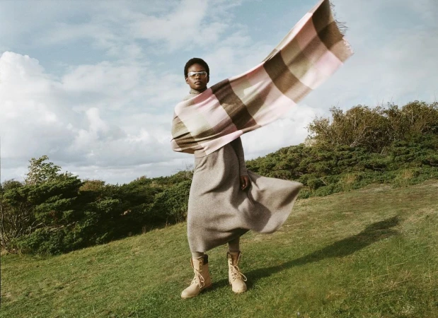 Adult woman wearing tan boots and a long grey dress standing in a grassy field with a pink and beige checkered scarf waving in the wind.