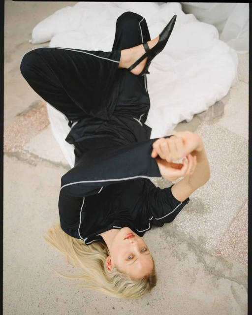 Blonde woman in black athletic wear poses on a marbled floor with white bedding, shot from above with focus on her face and attire.
