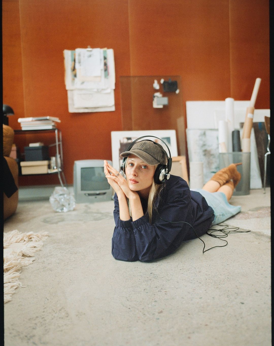 Young woman in headphones and cap lies on the floor in a room with neutral colours, vintage TV, and various objects in background.