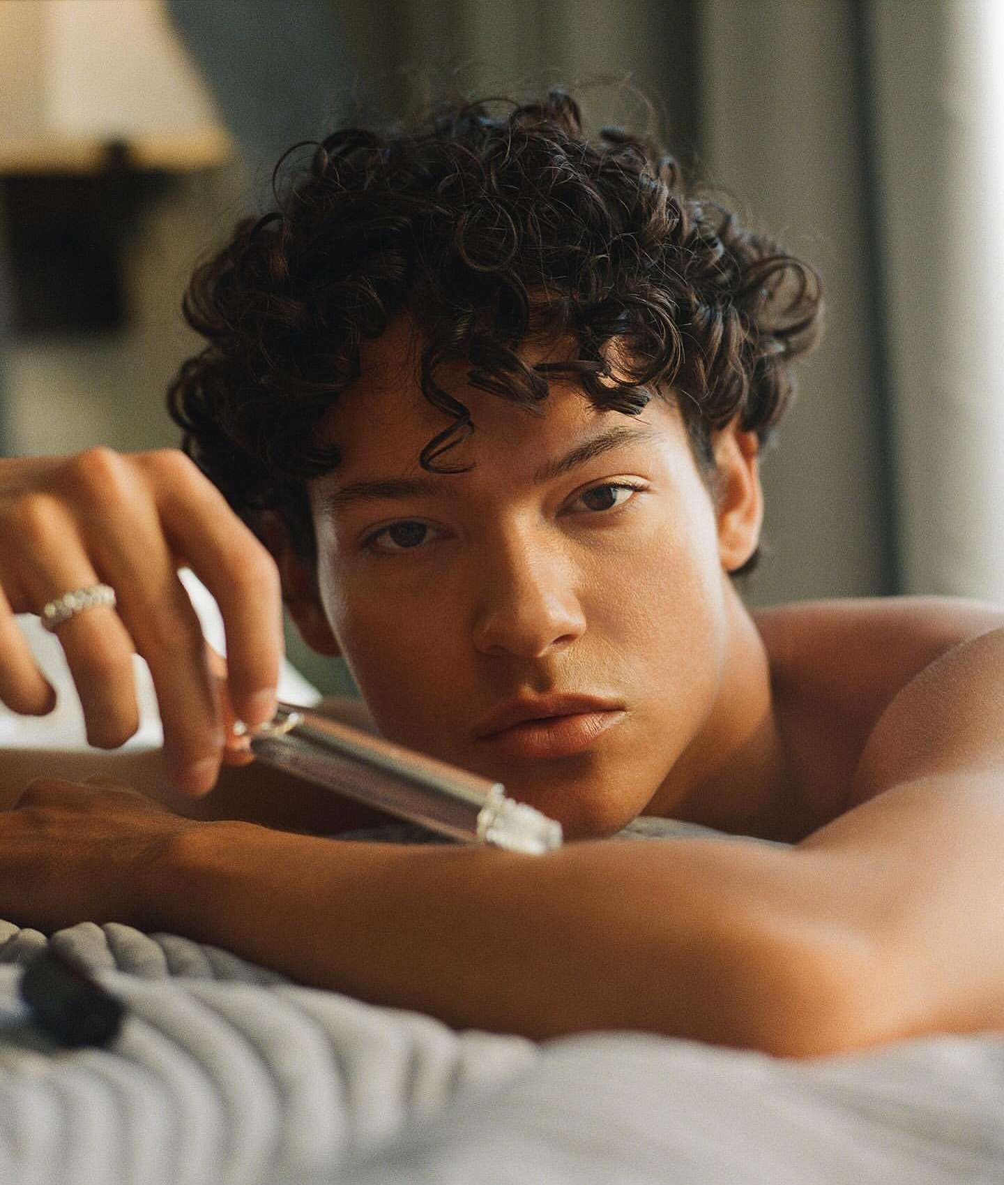 Attractive young man with curly hair, wearing a ring, and holding a silver object while laying on a textured grey surface indoors.