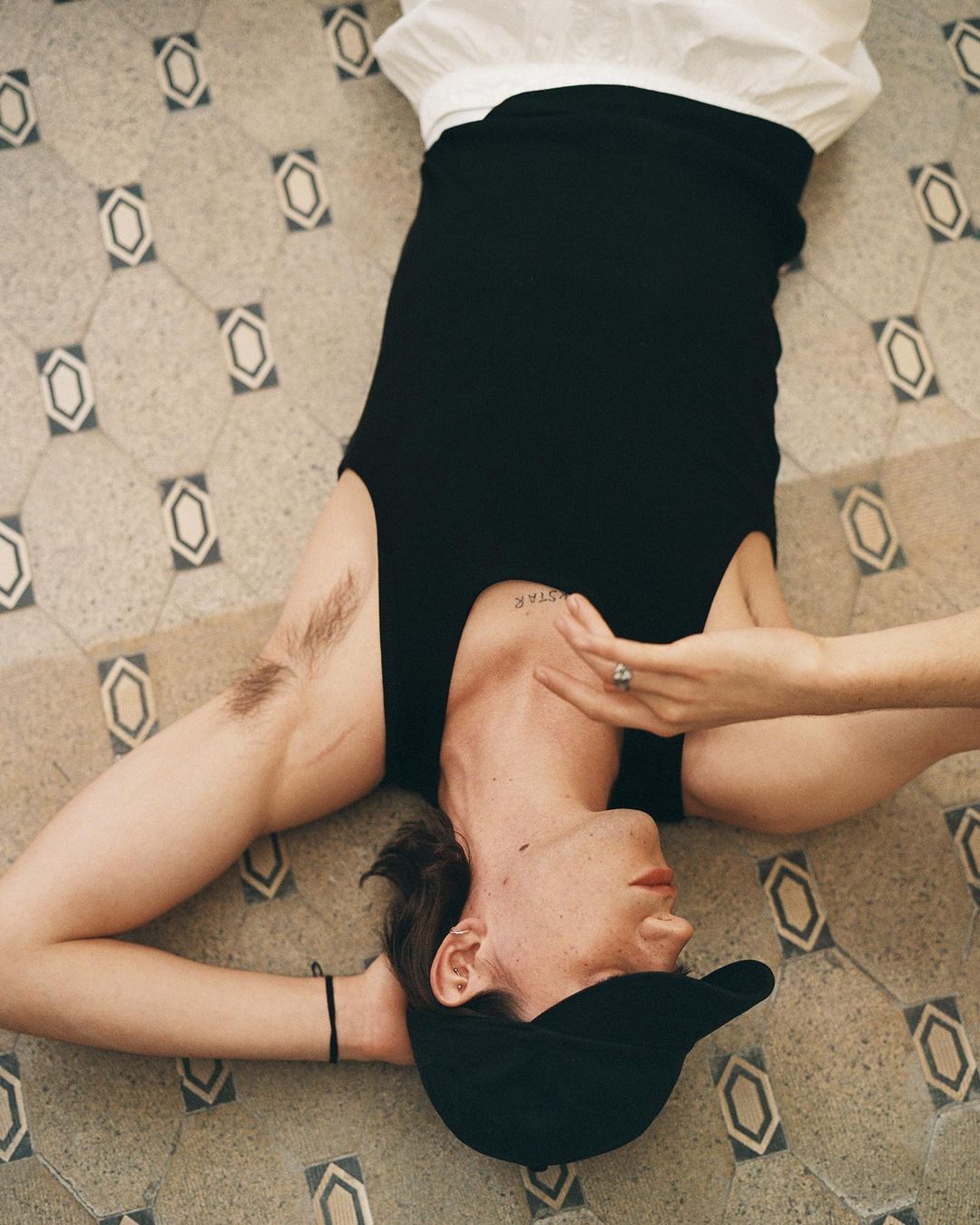Young adult lies on patterned tile floor wearing a black top and baseball cap. Focus is on the person with neutral expression.