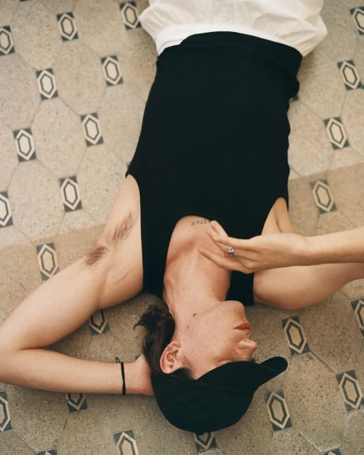 Young adult lies on patterned tile floor wearing a black top and baseball cap. Focus is on the person with neutral expression.