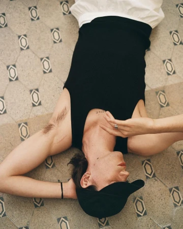 Young adult lies on patterned tile floor wearing a black top and baseball cap. Focus is on the person with neutral expression.