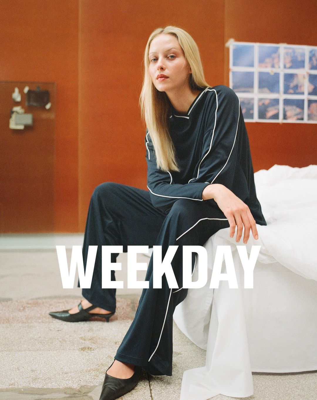 A woman sits in black pants and a long-sleeve top on a white bed with a rust-colored wall in the background with a white logo.