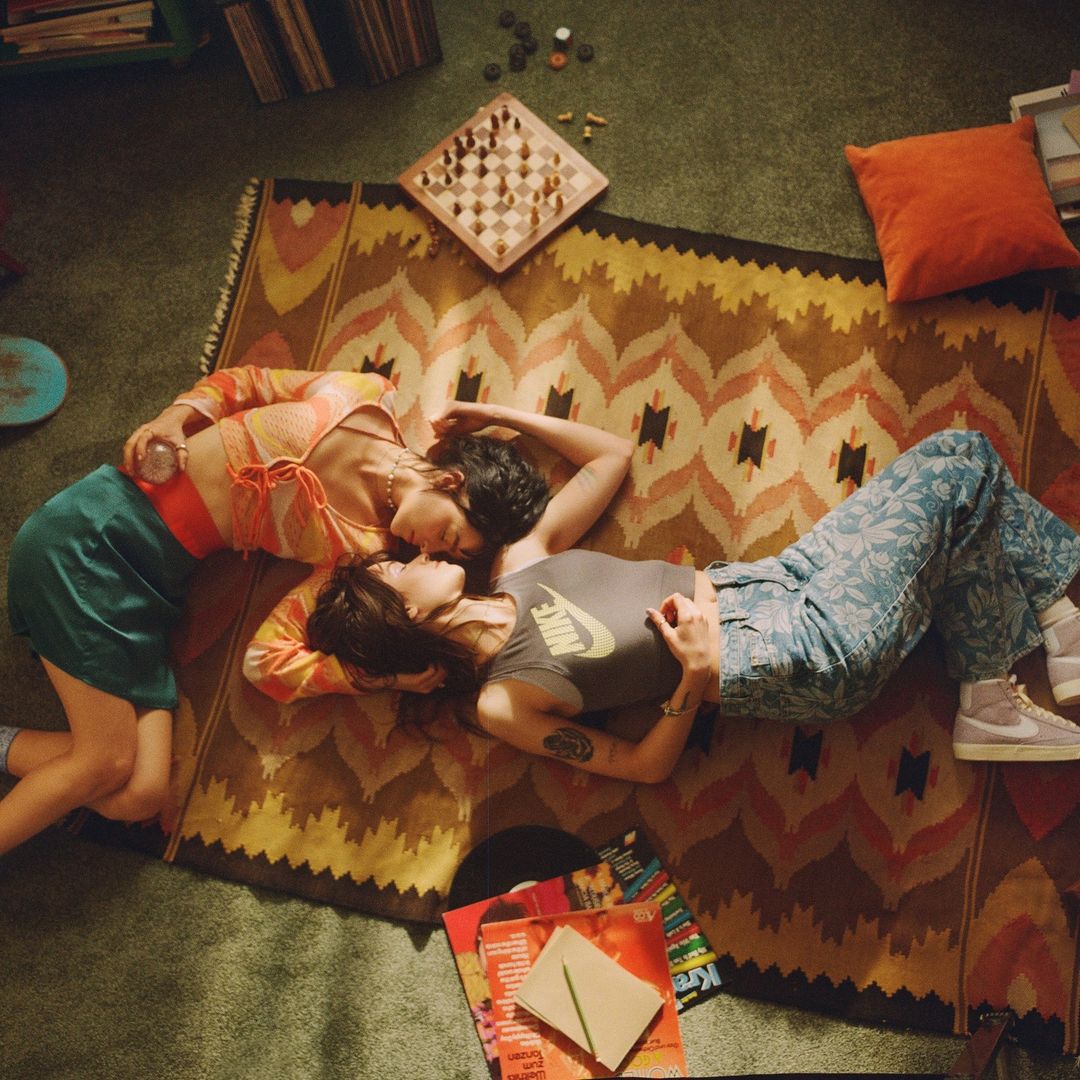 Two women lie face-to-face on a patterned rug with records and a chessboard, creating a warm, intimate indoor lifestyle image.