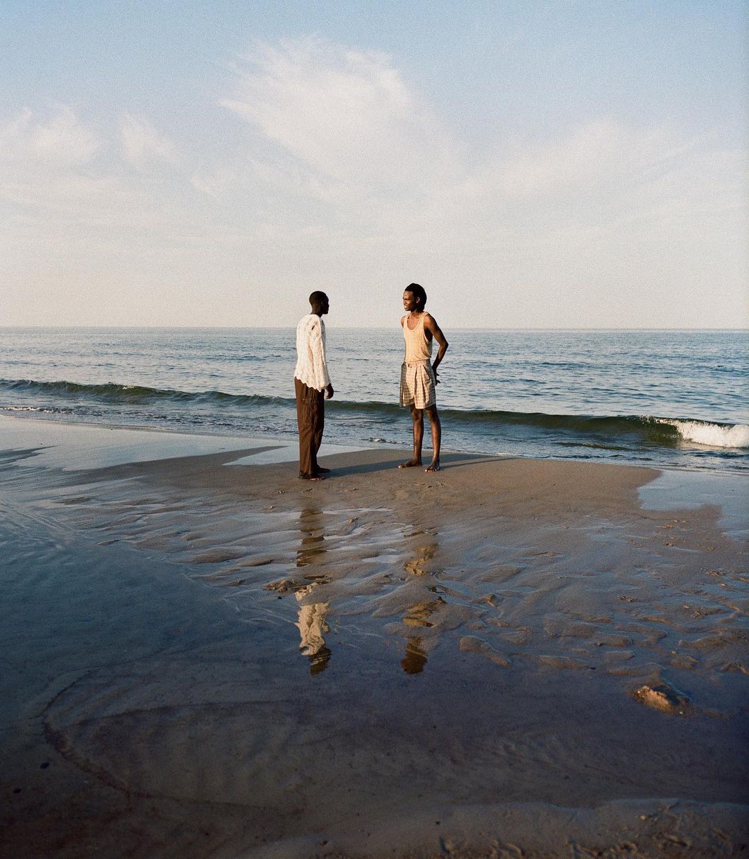 Two African men stand on a wet sand beach, facing each other near the water's edge, with reflections mirrored in the damp sand.