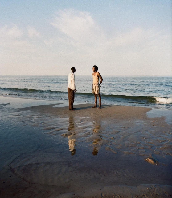 Two African men stand on a wet sand beach, facing each other near the water's edge, with reflections mirrored in the damp sand.