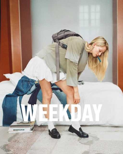 Blonde woman in casual fashion bending over bed with jeans, backpack, and books, shot indoors with natural lighting. 