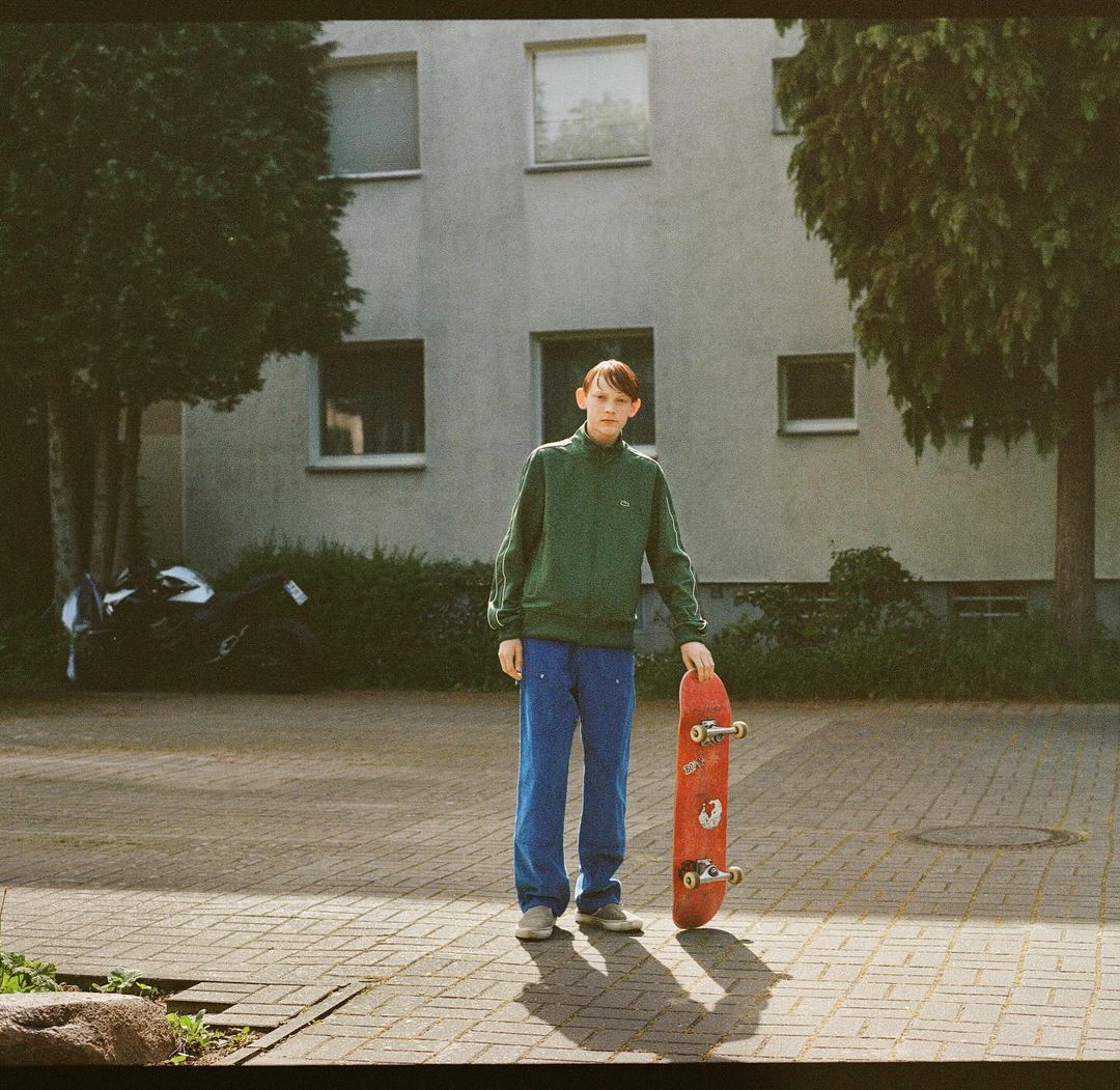 Teenager in a green jacket poses outdoors holding a skateboard, with an apartment building and a parked motorcycle in the background.