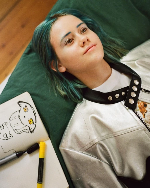 Teen with dyed blue hair and silver jacket lies on green cushion with sketchpad and markers.  Neutral expression, studio lighting.