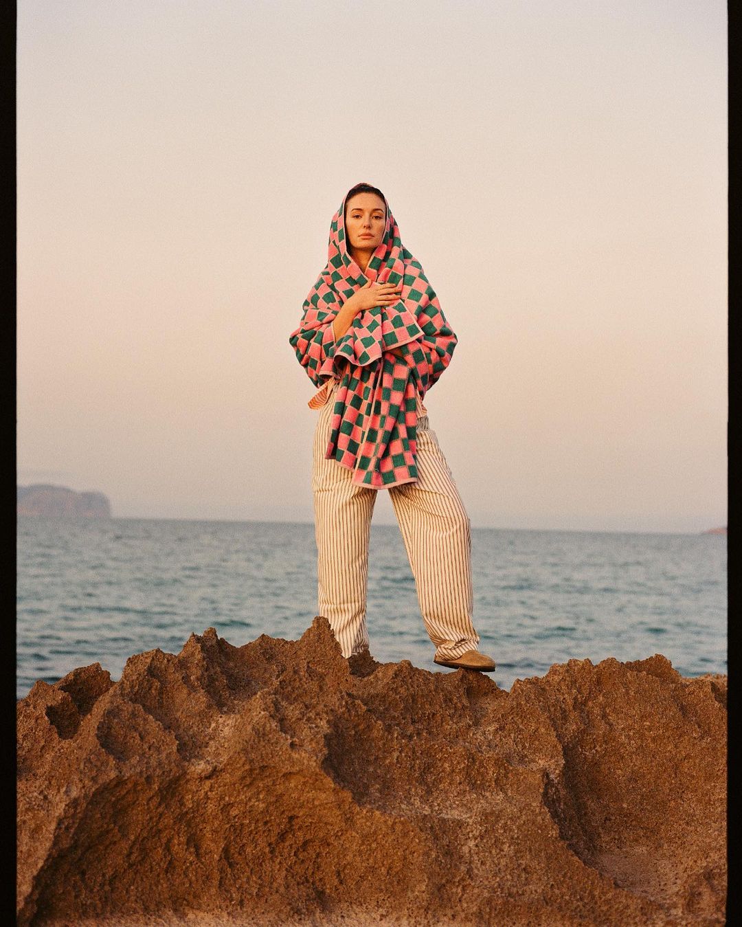 Adult woman stands on rocks wearing striped pants and draped in a checkered shawl, overlooking a serene seascape at dusk.
