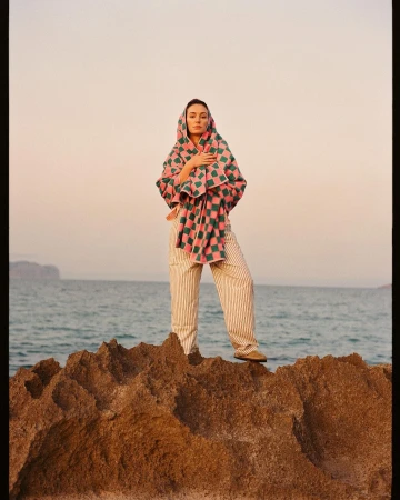Adult woman stands on rocks wearing striped pants and draped in a checkered shawl, overlooking a serene seascape at dusk.