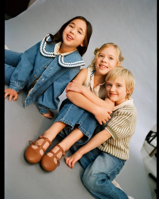 Three smiling children pose in a studio setting; one wears a denim jacket, another a striped top, and the third embraces them.