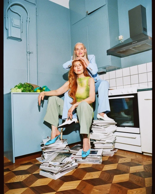 Two women in casual suits sit on magazines in a kitchen, styled with pale blue walls, tiles, and cabinets, and an angular parquet floor.