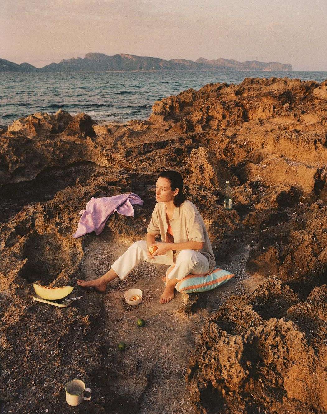 Woman relaxes on rocky shore near ocean with cut melon, limes, and water bottle at sunset, wearing casual clothing and sitting barefoot.