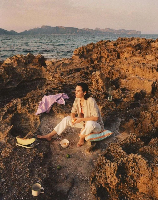 Woman relaxes on rocky shore near ocean with cut melon, limes, and water bottle at sunset, wearing casual clothing and sitting barefoot.
