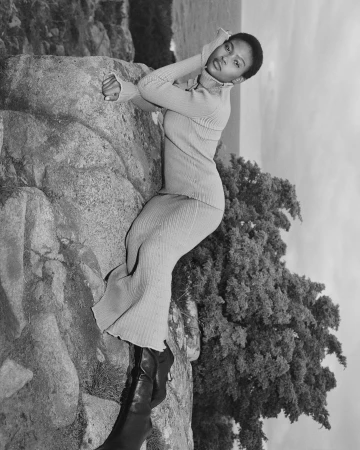 Black woman wearing a ribbed turtleneck sweater and long skirt leaning against rock formations outdoors.