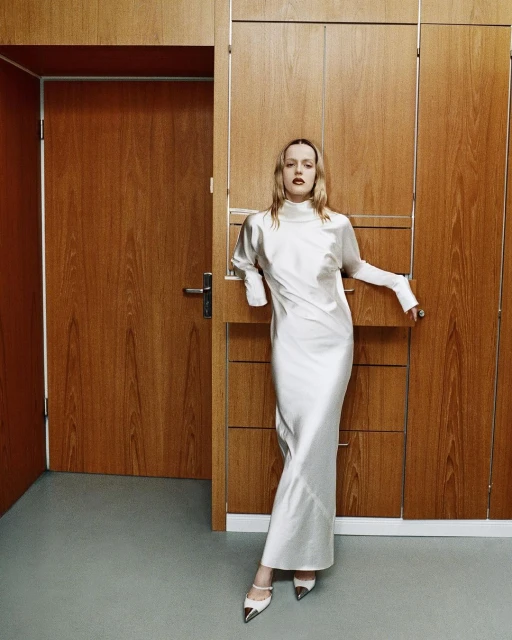 A slender adult woman in a stylish white dress and matching shoes stands against a background of wooden doors and light grey flooring.