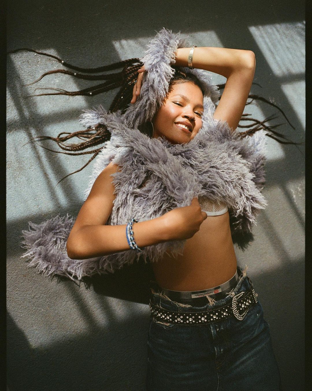 Smiling woman in jeans and faux-fur jacket lies on a grey surface with sunlight and shadows, posing with her hand near her head.