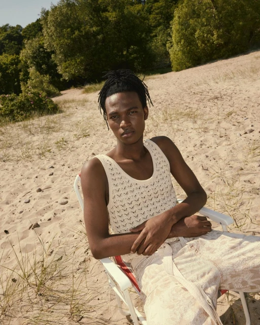 Portrait of a Black male model sitting in a beach chair on a sandy dune, wearing a tank top and loose pants with trees in the background.