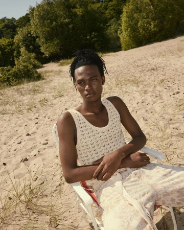 Portrait of a Black male model sitting in a beach chair on a sandy dune, wearing a tank top and loose pants with trees in the background.